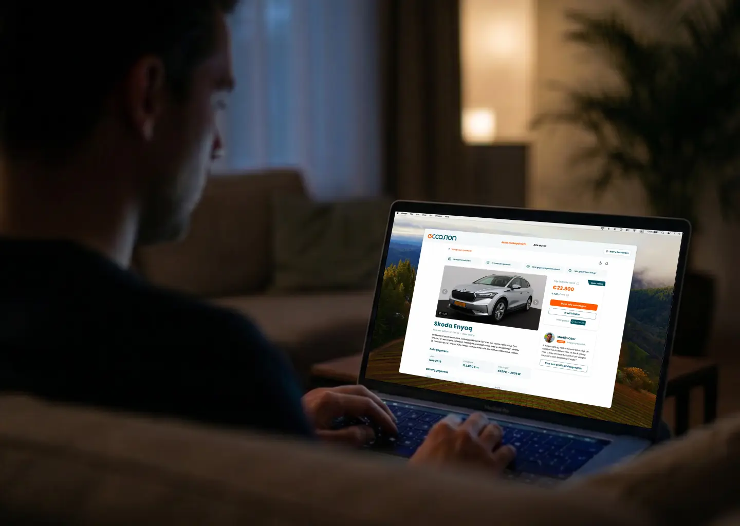 Someone on a sofa browsing the Eccasion car platform on a laptop, showing a Skoda Enyaq listing.